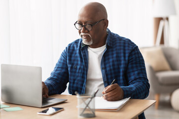 Senior Black Man Using Laptop And Writing Report Taking Notes Working Online Sitting At Desk In...