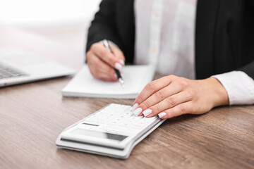 Banker with notebook using calculator at wooden table in office, closeup