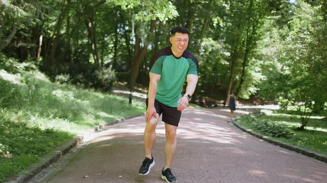Asian runner athlete with knee pain in park. Japanese man massaging stretching, trauma injury while jogging outdoors. Fitness male guy sprain severe pain stretch pull. Leg muscle cramp calf sport.
