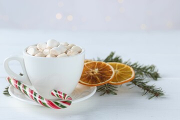 Yummy cocoa with marshmallows in cup, candy cane, dried orange slices and fir tree branches on white wooden table. Space for text
