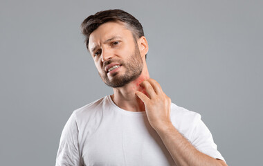 Middle aged bearded man scratching highlighted with red neck on grey studio background, copy space....
