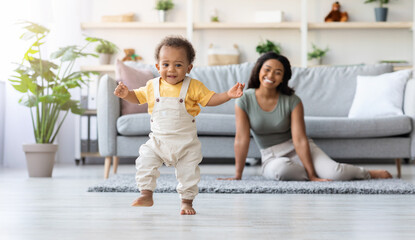 First Steps. Adorable Black Infant Child Walking In Living Room At Home, Cute African American Toddler Boy Stepping On Floor And Looking At Camera, His Proud Mother Smiling On Background, Free Space