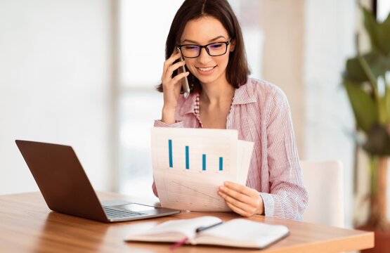 Checking Papers. Young businesswoman in glasses talking on mobile phone, looking at document with graphs, copyspace
