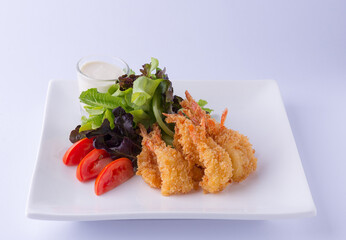 Fried shrimp salad with green lettuce, green oats, red tomatoes and white dressing on a white square plate on a white backdrop.