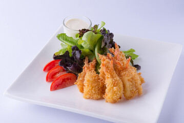 Fried shrimp salad with green lettuce, green oats, red tomatoes and white dressing on a white square plate on a white backdrop.