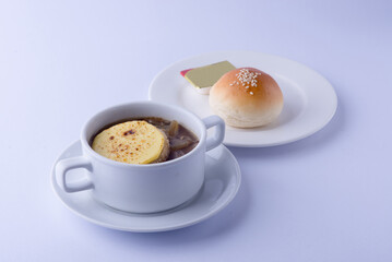 Clear onion soup in a white bowl, with sliced ​​potatoes, served with buttered bread on a white plate, on a white backdrop.