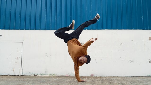 Hispanic man stretch arms and dance street dancing in front of wall. Motion shot of stylish dancer or choreographer in casual outfit practicing dancing in hip hop style. Outdoor sport 2024. Hiphop.