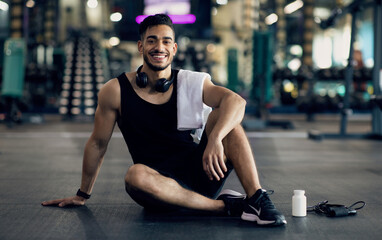 Young Arab Male Athlete Relaxing On Floor After Training At Gym, Smiling Middle Eastern Man Resting Next To Blank Container With Supplement Pills, Enjoying Bodybuilding And Healthy Lifestyle