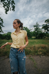 A young woman in a yellow shirt and ripped jeans stands outdoors, surrounded by greenery and under a cloudy sky.