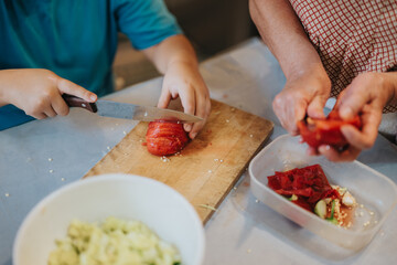 A child and adult work together in the kitchen, slicing fresh red tomatoes on a wooden cutting board. The image conveys teamwork, cooking skills, and family bonding.