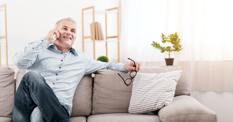 Communication Concept. Happy senior man calling his children, talking on mobile phone, resting on couch, copy space