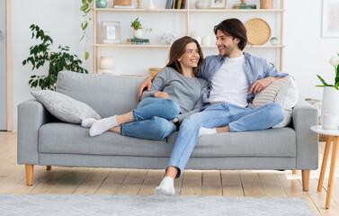 Fototapeta premium Young couple in love hugging on sofa at home, looking at each other, enpty space