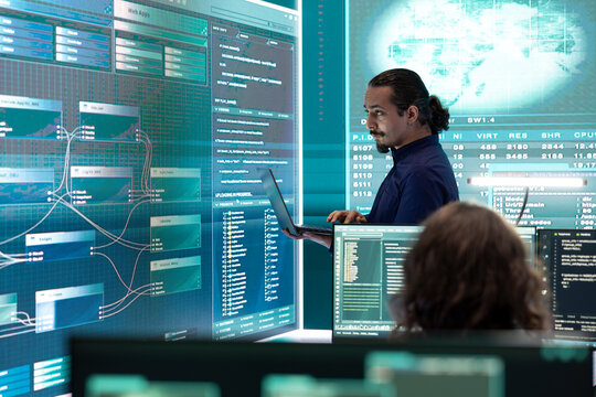 Governmental hacker monitoring cyber attacks on a big screen, compromising online security. Specialist infiltrating digital networks, breaching security protocols. Cyber surveillance activity.