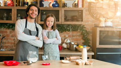 Young Handsome Dad And His Little Daughter In Aprons Posing In Kitchen While Preparing Pastry Together, Enjoying Homemade Food, Cooking Pie, Baking Sweets, Standing With Folded Arms, Smiling At Camera