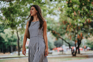 A joyful woman wearing a striped dress is strolling through a leafy park. The vibrant green background and sunshine create a relaxed and refreshing ambiance, perfect for a casual walk.