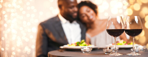 Selective focus on two glasses of wine over cuddling black couple, having festive dinner at restaurant
