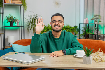 Hello. Indian smiling man smiling friendly at camera and waving hands gesturing hello hi, greeting or goodbye welcoming with hospitable expression at home office. Arabian guy sitting at table in room