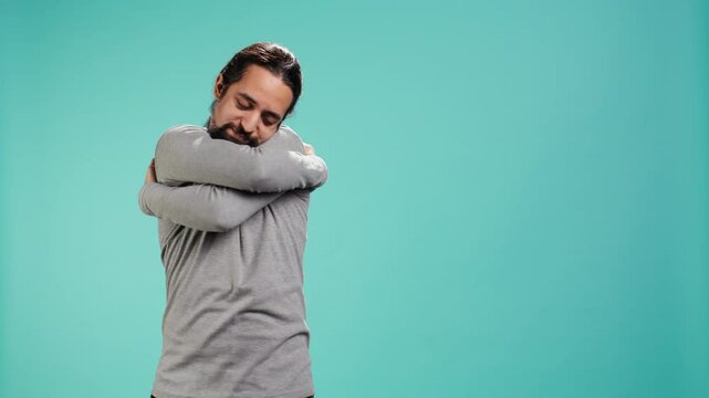 Portrait of man standing in studio, hugging himself in gesture of comfort and self love, isolated over studio background. Cheerful person doing self acceptance, embracing himself, camera B