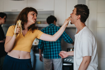 A group of young friends enjoying a playful moment in the kitchen. A woman feeds a man a piece of banana, while others cook in the background.