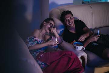 A happy couple relaxes together on a cozy sofa, sharing laughter and snacks in a warm evening...
