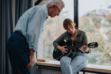 A teacher provides guidance to a student learning to play the acoustic guitar. The lesson takes place near a large window, creating a focused and peaceful atmosphere.