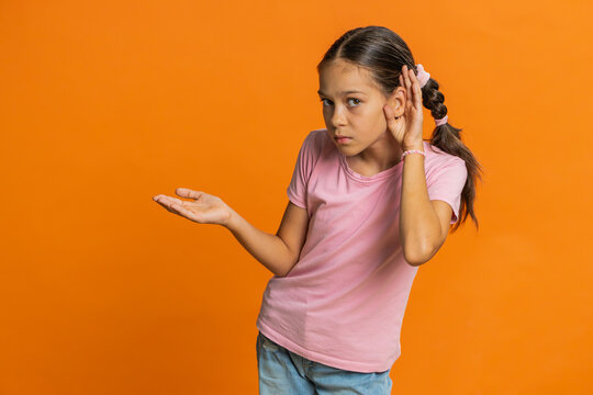 I cant hear you. What. Child girl kid trying hear you, looking confused and frowning, keeping arm near ear for louder voice asking to repeat to hear information deafness. Children on orange background