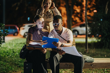 Young adults collaborating on a project at night, using a digital tablet. They are focused on work while a colleague talks on the phone, surrounded by evening city lights.