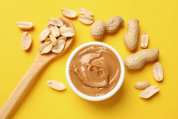 Tasty peanut butter in bowl and spoon with groundnuts on yellow table, flat lay