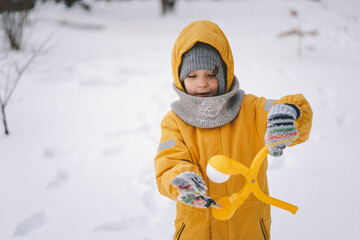 Young child wearing a bright yellow winter playing with a snowball maker in a snowy landscape...
