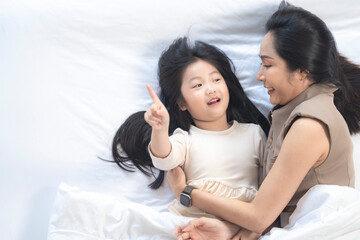 A joyful mother and daughter laughing together while lying on a bed. The playful moment showcases their strong bond and happiness in a comfortable home setting, surrounded by natural decor.