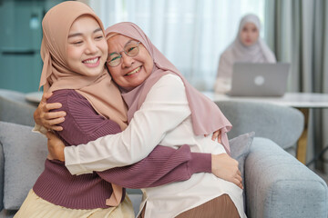 Obraz premium asian senior mother and adult daughter in hijab dresses sitting on sofa,embracing each other,smiling,background blurred a young arab woman working on laptop