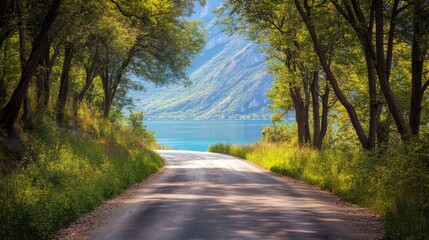 Obraz premium 2408 90.A quiet asphalt road meanders through a dense forest of green trees, with a clear blue lake visible just beyond the foliage. The mountains rise in the background, creating a dramatic