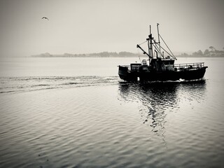 A lone fishing boat sails back in the calm afternoon waters. The misty horizon and a solitary bird in flight add to the tranquil scene.