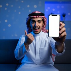 Young Arab muslim man in traditional clothes showing mobile phone isolated on home and sitting on sofa with blue colors.
