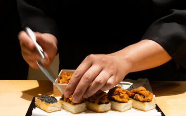 Close up of Japanese chef carefully plating sea urchin on toasted bread set showcasing fine dining preparation with precision and detail on a counter in the restaurant