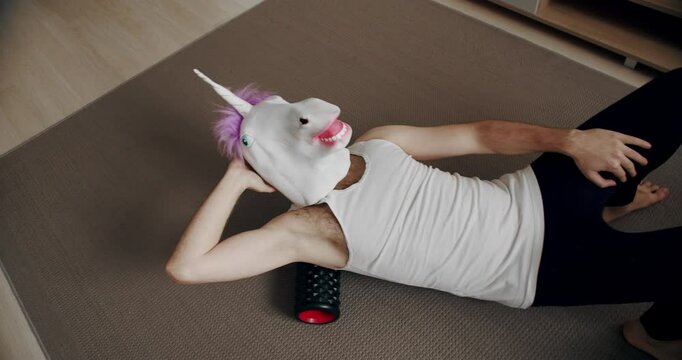 Funny man with unicorn mask doing back exercise with roller for myofascial relaxation at home. comical fitness at home. Foam roller for muscle and fascia stretching. Tools for self massage at home.