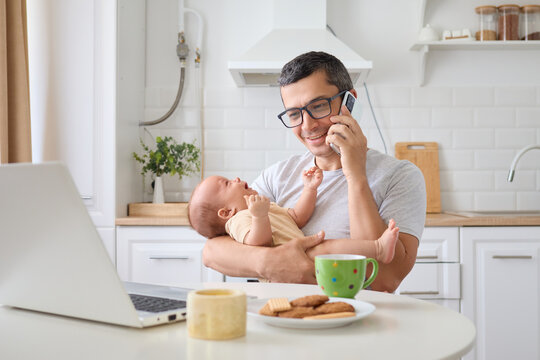 Cheerful father speaking on cell phone holding his newborn son during his remote workday on laptop in home kitchen interior