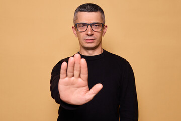 Bossy man wearing glasses and raglan standing indoors with serious expression holding up hand in...