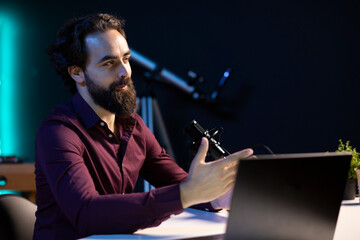 Media star sitting at table in dimly lit home studio interior, recording audiobook using professional microphone. Audio expert reading from laptop, doing recording for fans