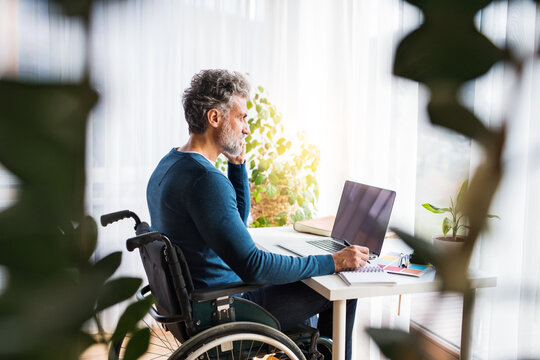 Handsome man in wheelchair working from home office, making phone call and writing notes in planner. Telecommuting and home office.