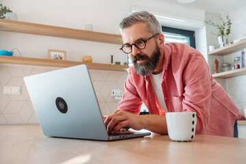 Mature man working from home on laptop, standing by kitchen island, with cup of coffee. Concept of...