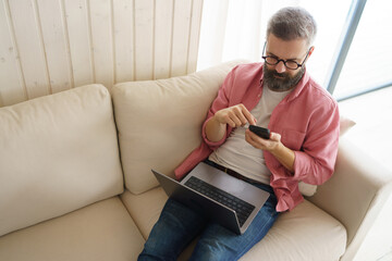 Fototapeta premium Man with notebook and smartphone sitting on sofa, online banking, internetbanking at home throught website and app. Paying with smartphone app.
