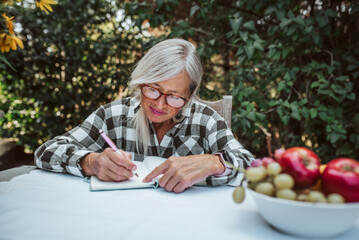 Beautiful older woman is sitting in the garden and writing in her diary. Female senior is keeping...
