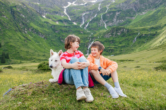 Happy brother and sister enjoying leisure time sitting with dog on meadow