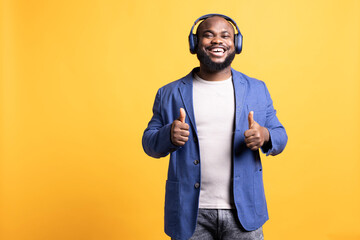 African american man having fun listening to music on headset, showing thumbs up sign. BIPOC person hearing tunes and showing positive recommendation hand gesturing, studio background