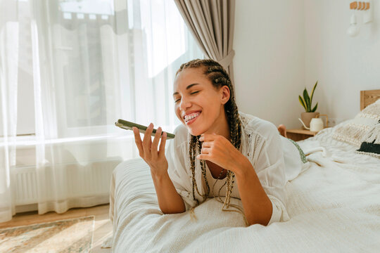 Happy woman sending voicemail through smart phone on bed at home