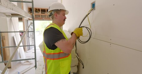 In a busy remodeling project, a hardworking construction worker, who is dressed in a safety vest and wearing a protective helmet, expertly manages and installs electrical wiring with precision