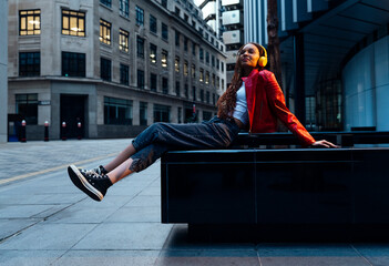 Thoughtful young woman listening to music though headphones on bench in city