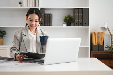 Successful businesswoman drinking hot beverage and reviewing documents at her desk