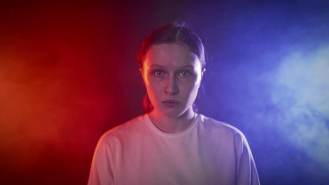 Young Caucasian woman with big eyes is afraid and looking at camera, portrait. Talented actress self-expressing, touching her body and head, slow motion in dramatic red and blue light, fear and horror